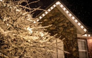 Snow-covered branches glisten under the soft light, with a stone-clad house adorned with holiday lights in the background. A wintery scene evoking a cozy, festive mood.