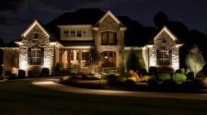 Landscape lighting illuminates the exterior and grounds of a stone house at night.
