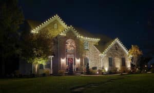 Brick home illuminated with warm white Christmas lights along the roofline. A festive wreath adorns the front window, enhancing the holiday cheer. Professional Christmas light installation creates a welcoming ambiance.