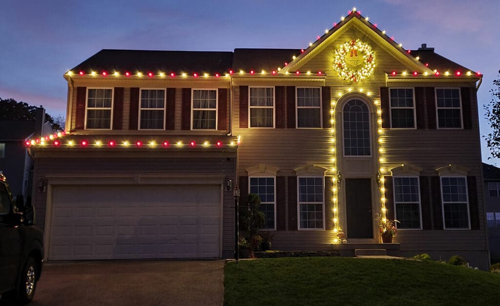 Two-story house decorated for Christmas with red and yellow lights outlining the roof and a large wreath above the front door.