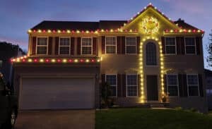 Two-story house decorated for Christmas with red and yellow lights outlining the roof and a large wreath above the front door.