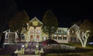 Large house decorated with white Christmas lights and wreaths. Lights adorn the roof, trees, and stone walls, creating a festive holiday scene at night.