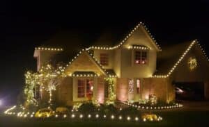 A house is decorated with warm white Christmas lights outlining the roof, bushes, and a small tree, creating a cozy and festive holiday scene at night.
