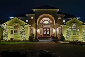 Elegant home with Christmas light installation: Conical trees adorned with warm white lights flank the grand entrance, which is also decorated with wreaths and pumpkins. The driveway leads up to the illuminated doorway.
