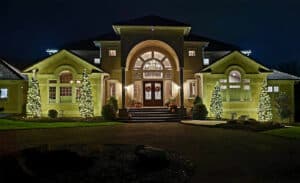 Elegant home at night with professional Christmas light installation. Two lit Christmas trees flank the entrance, wreaths adorn the double doors, and subtle lighting highlights the architecture.