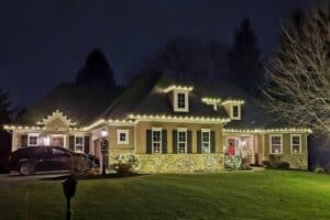 Warm white Christmas lights outline a house with dormer windows and illuminate shrubs beside the entrance.