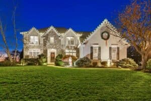 White Christmas lights trace the gabled roof of a stone and stucco house, with a wreath above the garage.