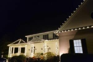 Warm white Christmas lights trace the roofline and wrap the porch columns of a two-story house.