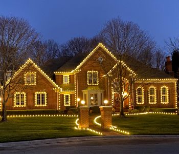 Warm white Christmas lights outline a brick house, its windows and the curved walkway to the entrance.