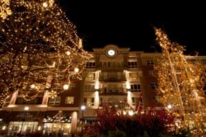 Festive commercial Christmas lighting illuminates a brick building with a clock tower. Trees sparkle with warm white lights, creating a magical holiday atmosphere in the city at night. And-Sons Jewelers storefront is visible.