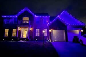 Purple Christmas lights outline the roofline of a two-story brick house at night.