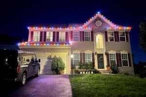 Multicolored Christmas bulbs trace the roofline of a two-story house at night.