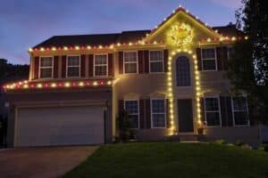 Red and warm white Christmas lights outline a two-story house, with a lit wreath above the arched entrance.