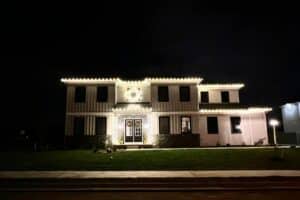 White Christmas lights outline a two-story house, with a lit wreath above the illuminated entrance.