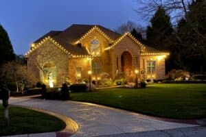 Warm white Christmas lights outline three roof peaks of a brick house beside a curved driveway.