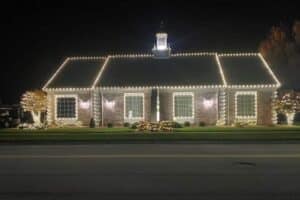 White Christmas lights outline the roof and windows of a brick building, with illuminated trees on either side.