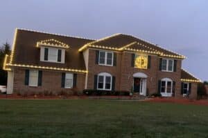Warm white Christmas lights outline the roof edges of a large brick house, with a lit wreath above the entrance.