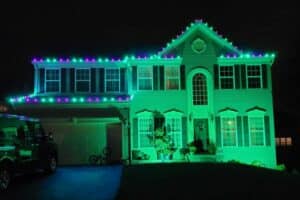 Green and purple Christmas lights outline the roofline of a two-story house at night.