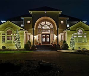 White Christmas lights wrap four trees flanking a house with a large arched entrance.