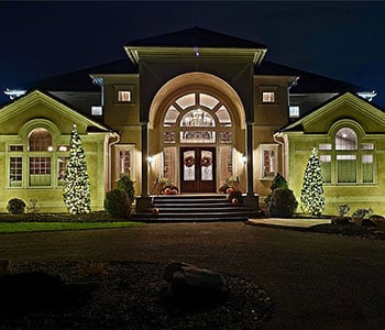White Christmas lights wrap four trees flanking a house with a large arched entrance.