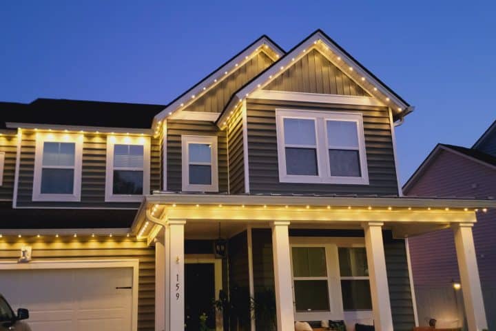 Warm white lights outline the roofline and porch of a gray two-story house at dusk.