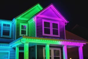 Green, purple and blue lights illuminate the roof peaks and porch of a two-story house at night.
