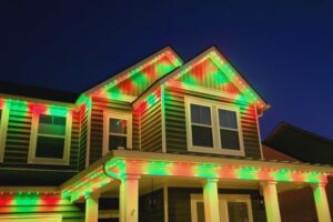 Red and green lights illuminate the roofline and porch of a gray two-story house at dusk.
