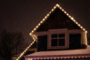 Warm white lights outline a steep roof peak above an upstairs window on a snowy night.