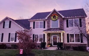 Festive two-story house decorated with Christmas lights and a wreath. The home features a stone facade and siding, with an Alabama "A" flag in the yard, set against a beautiful twilight sky.