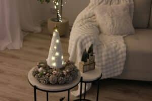 A cozy, small space holiday decor scene features a lit, yarn-wrapped Christmas tree centerpiece on a table, surrounded by a pinecone wreath. A fluffy white blanket adorns a couch in the background, enhancing the warm, festive mood.