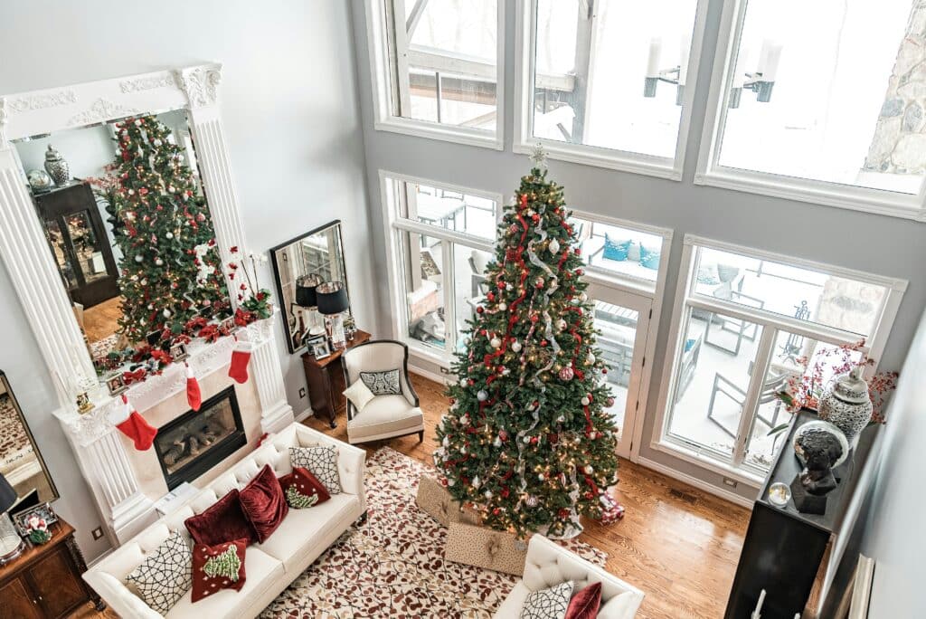 A bright, airy living room showcases winter living room decor, featuring a tall Christmas tree, white sofa with festive pillows, and a white fireplace mantel adorned with stockings and garland.