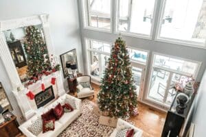 A bright, airy living room showcases winter living room decor, featuring a tall Christmas tree, white sofa with festive pillows, and a white fireplace mantel adorned with stockings and garland.