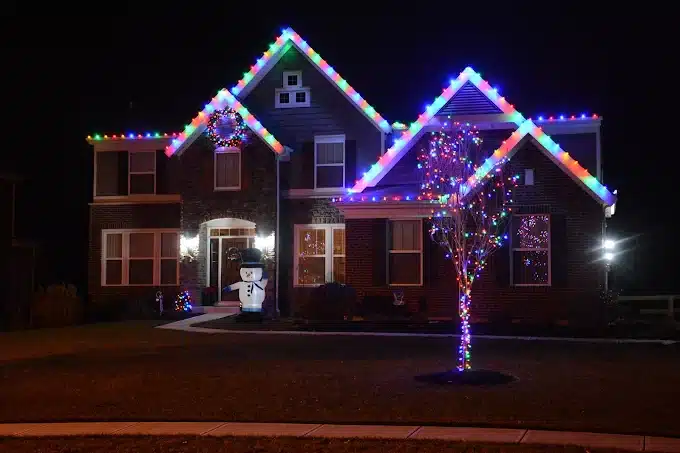 Festive two-story home at night, decorated with colorful Christmas lights outlining the roof and windows. A lit snowman stands on the lawn, adding to the cheerful holiday scene.
