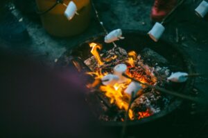 Marshmallows roasting over a crackling fire in a DIY home fire pit, creating a cozy evening atmosphere. Sticks with marshmallows are held over the flames, with a bucket nearby.