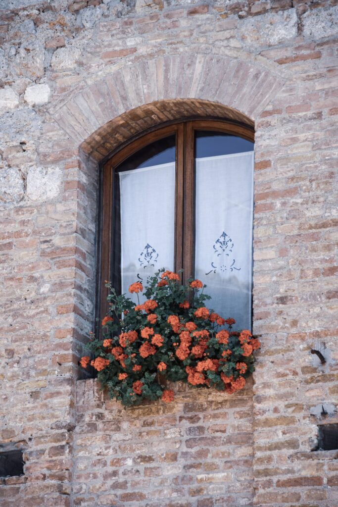 Arched window in a brick wall adorned with a vibrant orange flower box. White curtains with blue embroidery hang inside, adding charm to this DIY home window box installation.