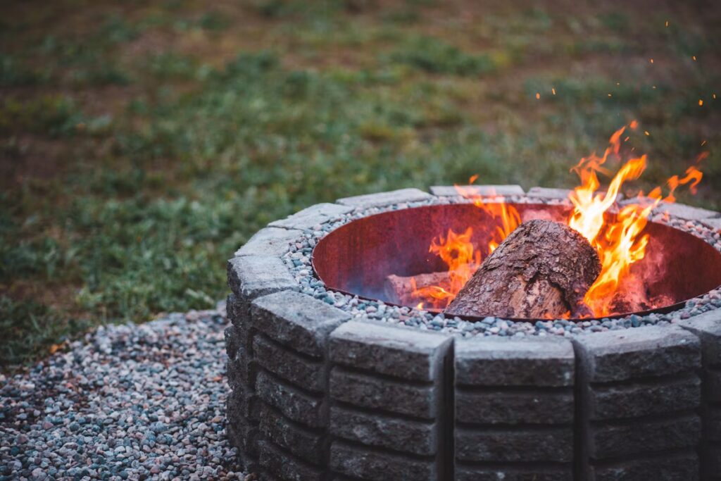A log burns in a circular stone fire pit surrounded by gravel and grass.