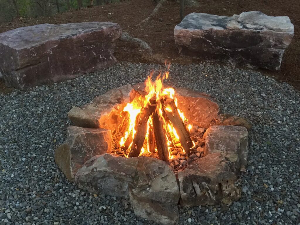 A wood fire burns inside a ring of stones on gravel, with large boulders nearby.