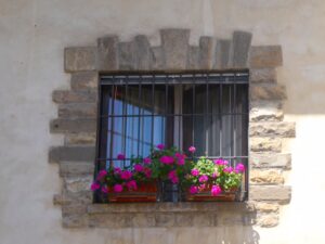 Stone window with iron bars and vibrant pink geraniums in window boxes. A charming DIY home window box installation adds seasonal charm with its colorful display.