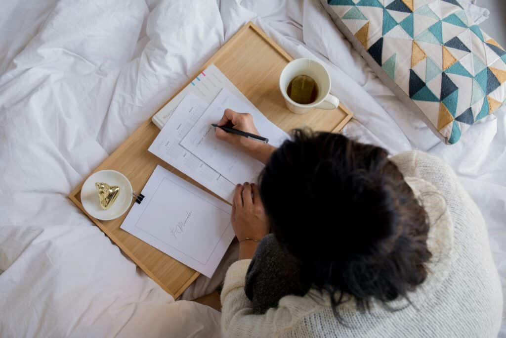 Woman in bed with a Christmas checklist on a wooden tray, writing with a pen. Mug of tea, decorative pillow, and small trinket dish also on the bed.