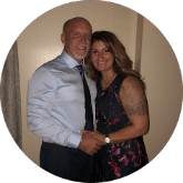 A smiling couple poses indoors; the man in a blue dress shirt and black tie, the woman in a sleeveless floral dress with a wristwatch.