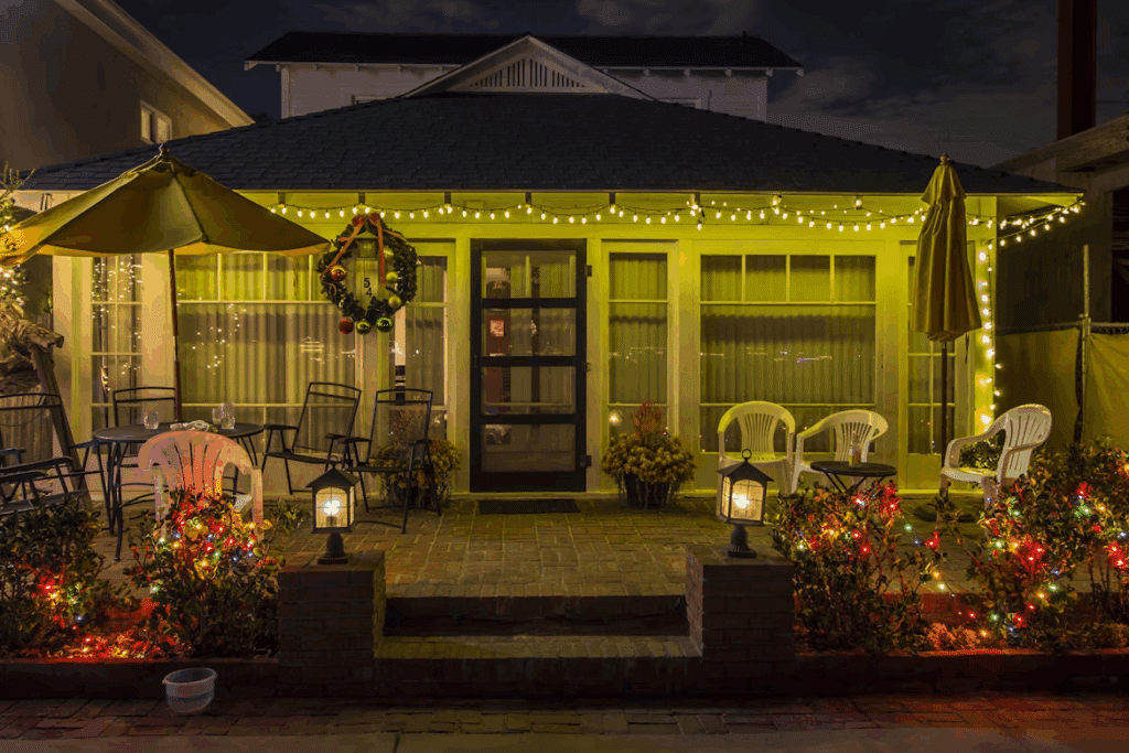 Cozy home exterior decorated with holiday lights and a wreath. Yellow string lights illuminate the porch, while colorful lights adorn bushes, creating a festive ambiance. Patio furniture and umbrellas add to the welcoming scene.