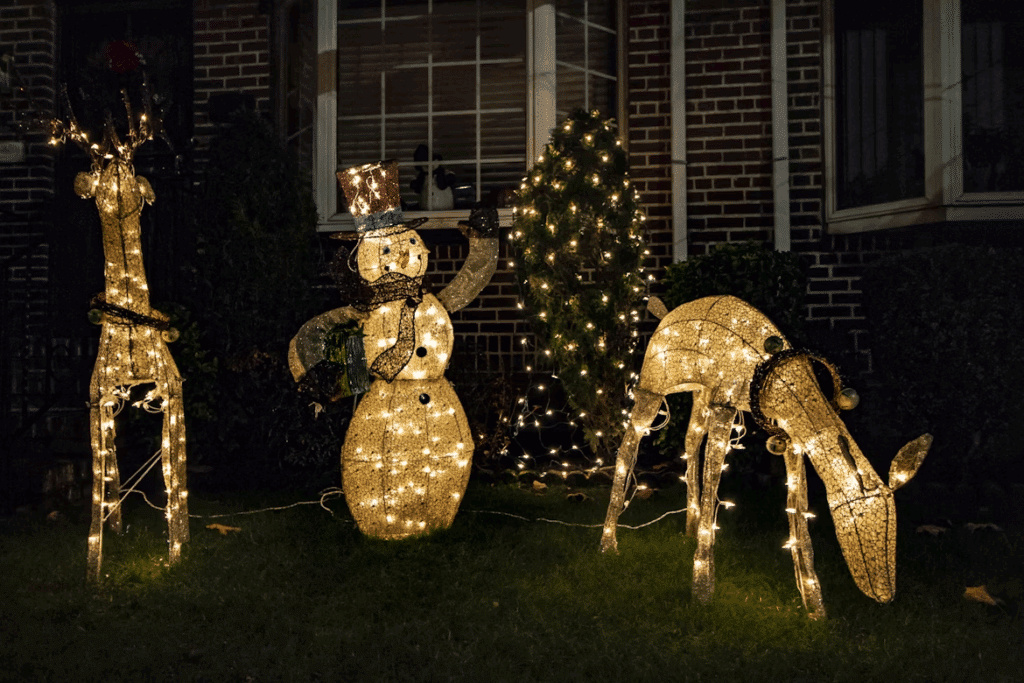 Festive holiday lights display with two illuminated reindeer and a snowman on a lawn in front of a brick house. The warm glow of the lights creates a cheerful, seasonal atmosphere.