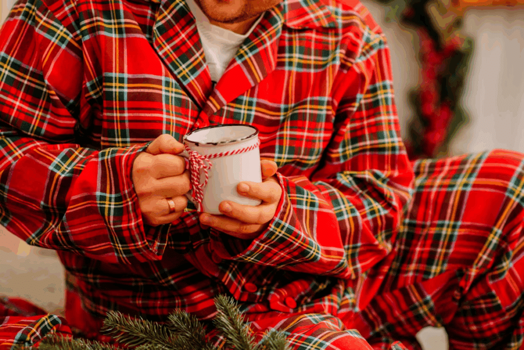 Man in red plaid Christmas pajamas holds a mug with a twine bow. Part of a Christmas checklist for a cozy, stress-free holiday.