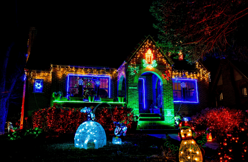 A brick house glows with vibrant Christmas lights: red bushes, blue window outlines, green trim, and a candle on the roof, illustrating festive holiday decor.