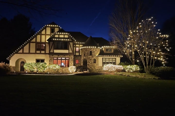 Elegant Tudor-style home at night, adorned with warm white Christmas lights outlining the roof and illuminating the landscaping. A stone turret adds to the fairytale charm.