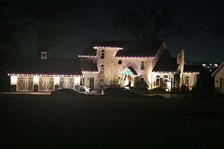 Night view of a large stone house decorated with red and white Christmas lights outlining the roof, creating a festive, warm glow. Garage doors are illuminated, adding to the holiday charm.
