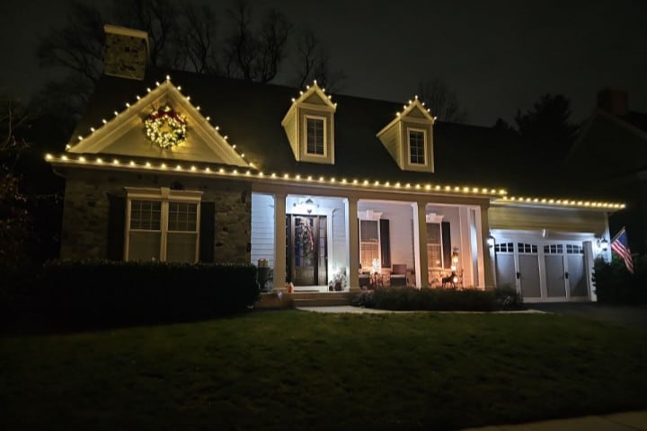 A charming house is adorned with warm white Christmas lights along the roofline and dormers, with a festive wreath above the front window. A covered porch and American flag add to the inviting holiday scene.