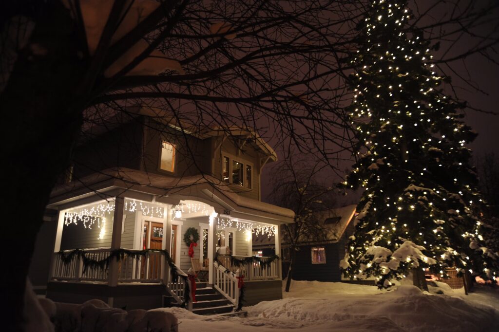 White icicle lights hang from a snow-covered porch beside a tall evergreen tree wrapped in Christmas lights.