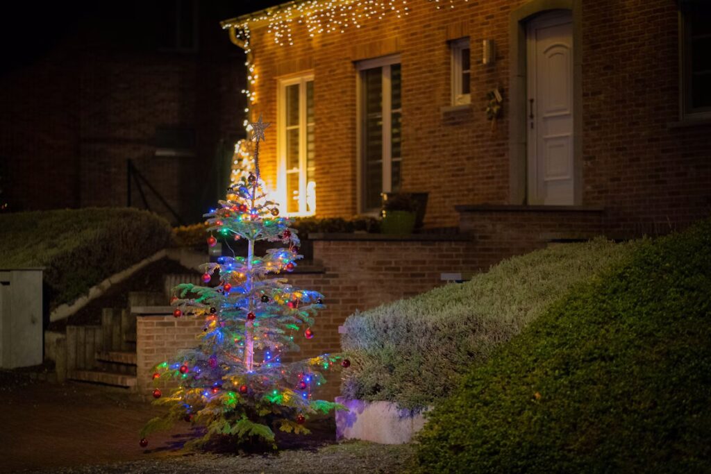 Outdoor Christmas lights adorn a brick house, with a decorated Christmas tree in the yard. The festive holiday display creates a warm, magical glow.