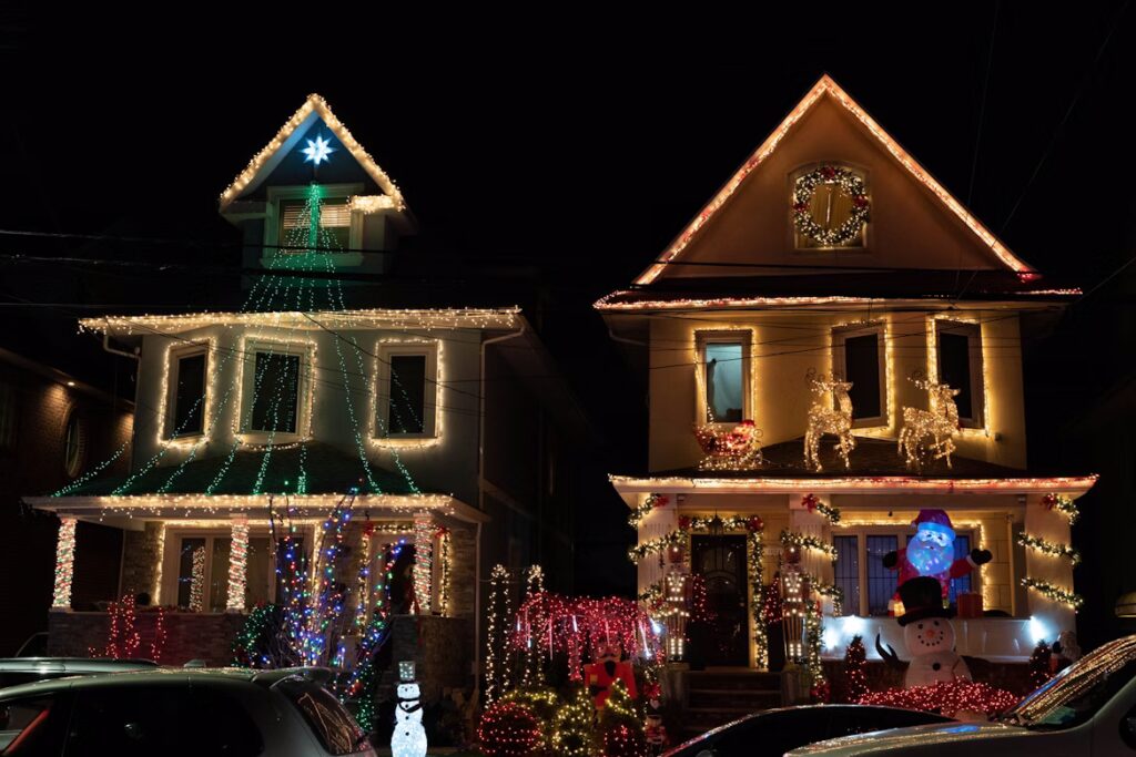Two houses decked out with Christmas lights create a dazzling holiday display. One house features cascading green lights and a snowman, while the other glows with warm white lights and reindeer.